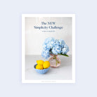 A magazine cover with light blue text says, The NEW Simplicity Challenge: 31 days to a simplified life. Below it, a pink vase of blue hydrangeas sits alongside a small blue bowl of yellow lemons on a white surface. Check out the Simplicity Challenge - E-Book by Simplified for more details!.