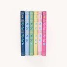 The Simplified Linen Journal Bundle features six vertically arranged journals with decorative floral spines. From left to right, the colors are dark blue, light blue, green, yellow, pink, and red. Each spine showcases a unique floral pattern that invites creativity and reflection.