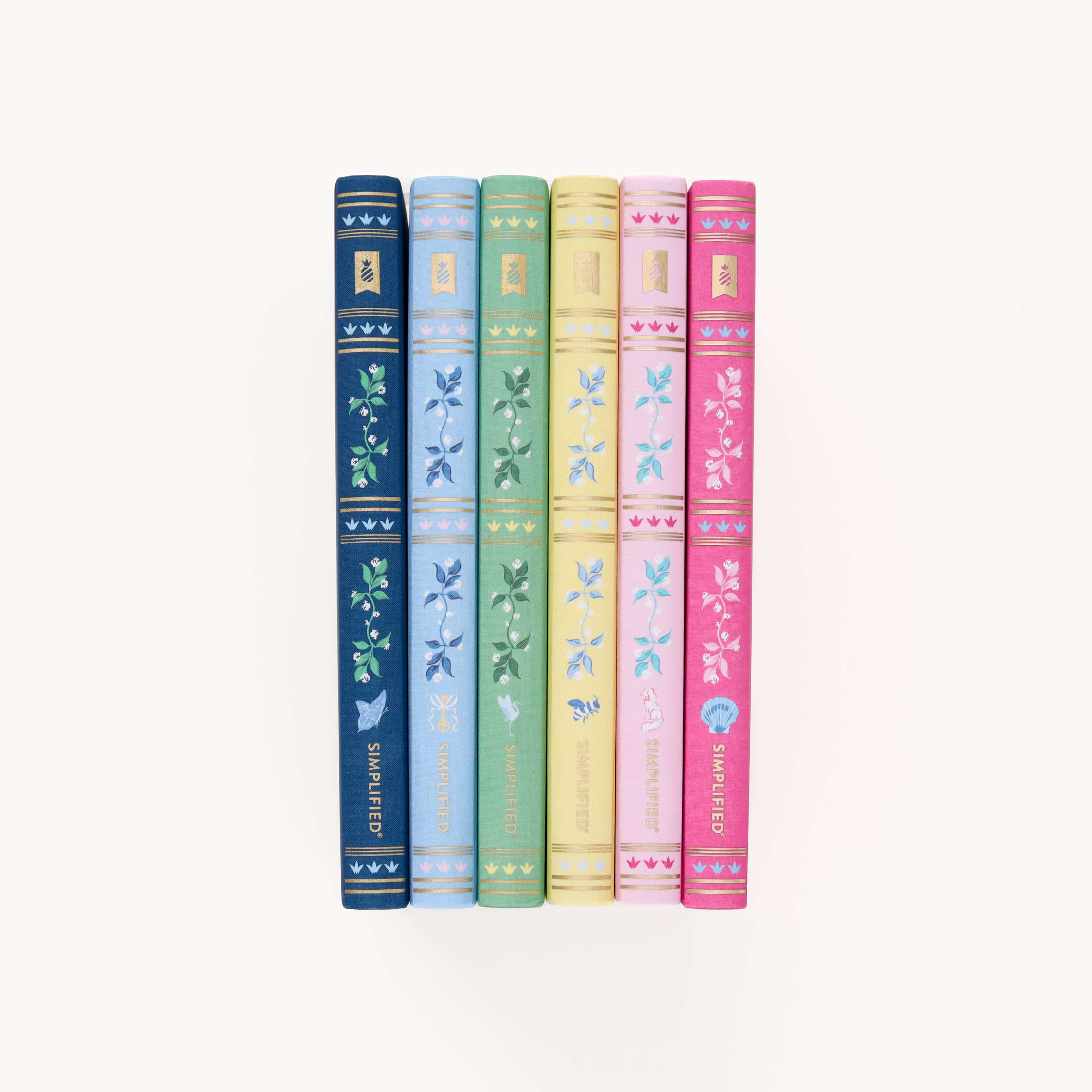 The Simplified Linen Journal Bundle features six vertically arranged journals with decorative floral spines. From left to right, the colors are dark blue, light blue, green, yellow, pink, and red. Each spine showcases a unique floral pattern that invites creativity and reflection.