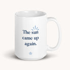 The Simplified ceramic mug, The Sun Came Up Again, boasts a positive blue print with a sunburst graphic above and a decorative emblem below, all set against a light background.